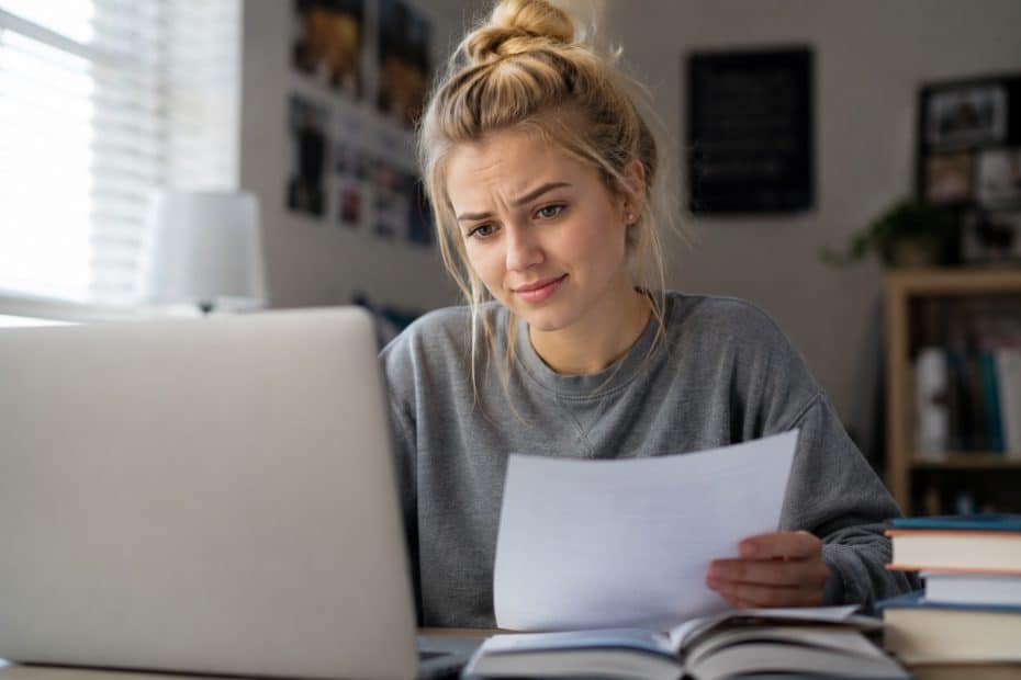 Young woman studying at a desk with a laptop, holding a paper and looking thoughtful.
